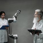 Dois pesquisadores de jaleco trabalham com um braço robótico em um laboratório.