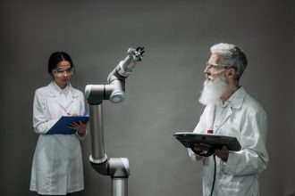 Dois pesquisadores de jaleco trabalham com um braço robótico em um laboratório.