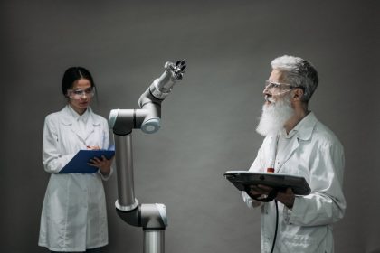 Dois pesquisadores de jaleco trabalham com um braço robótico em um laboratório.