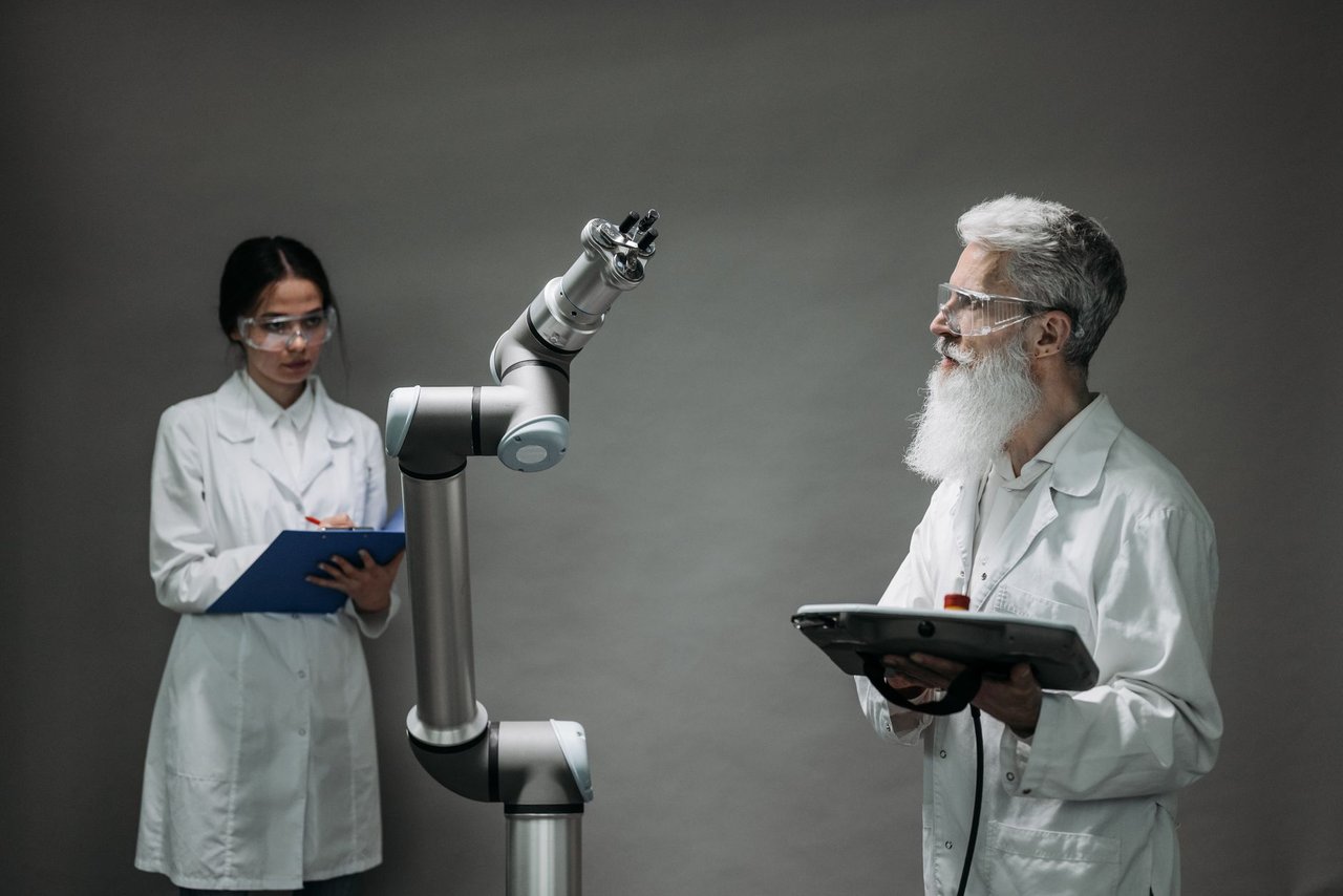Dois pesquisadores de jaleco trabalham com um braço robótico em um laboratório.