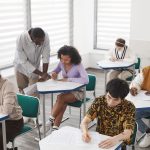 Grupo diverso de estudantes realiza uma prova em uma sala de aula, sob a supervisão de um professor.