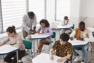 Grupo diverso de estudantes realiza uma prova em uma sala de aula, sob a supervisão de um professor.
