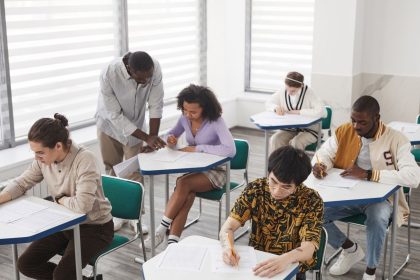 Grupo diverso de estudantes realiza uma prova em uma sala de aula, sob a supervisão de um professor.