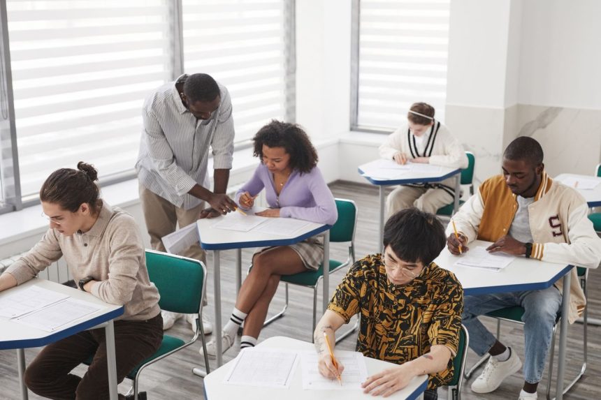 Grupo diverso de estudantes realiza uma prova em uma sala de aula, sob a supervisão de um professor.