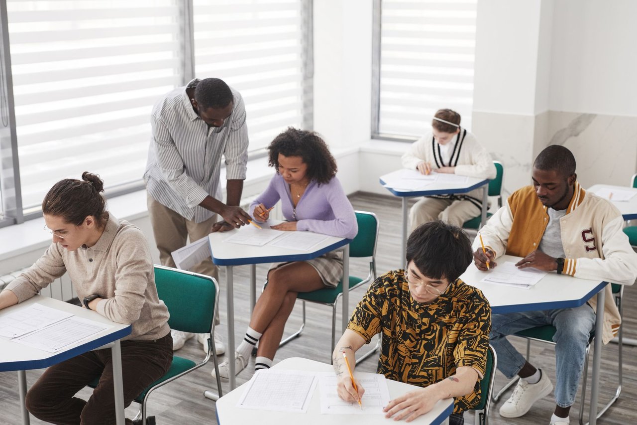 Grupo diverso de estudantes realiza uma prova em uma sala de aula, sob a supervisão de um professor.